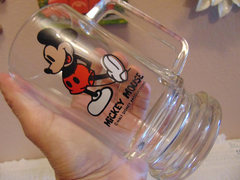 May include: A clear glass mug featuring a graphic of Mickey Mouse in red shorts and yellow shoes. The text MICKEY MOUSE and WALT DISNEY PRODUCTIONS is printed in black. The mug has a thick handle and a ribbed base design for a sturdy grip.