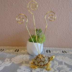 May include: A gold-toned turtle figurine featuring a shell encrusted with clear rhinestones and three tall, thin gold wire stems extending upward, each ending in a spiral loop designed to hold small cards or notes in an upright position.