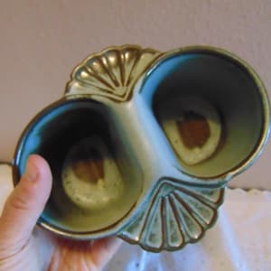 May include: A ceramic divided dish with two circular compartments and a decorative, fan-shaped edge. The dish features a blend of green, blue, and brown hues, with a glossy finish. The dish is held by a hand.
