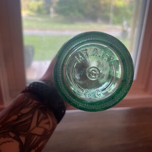 May include: A close-up of the bottom of a green glass bottle. The text "MT. AIRY" is embossed in a circular pattern, with "N.C." below. The bottle is held against a blurred background of a window and outdoor scenery.