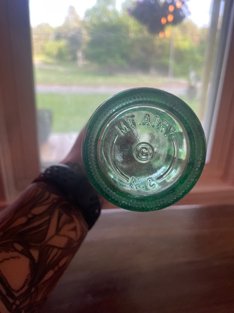 May include: A close-up of the bottom of a green glass bottle. The text "MT. AIRY" is embossed in a circular pattern, with "N.C." below. The bottle is held against a blurred background of a window and outdoor scenery.