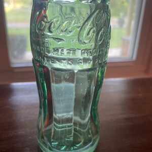 May include: A vintage green glass Coca-Cola bottle. The bottle has the Coca-Cola logo and other text embossed on the surface. The bottle is empty and has a classic, curved shape. The bottle is sitting on a dark brown surface.