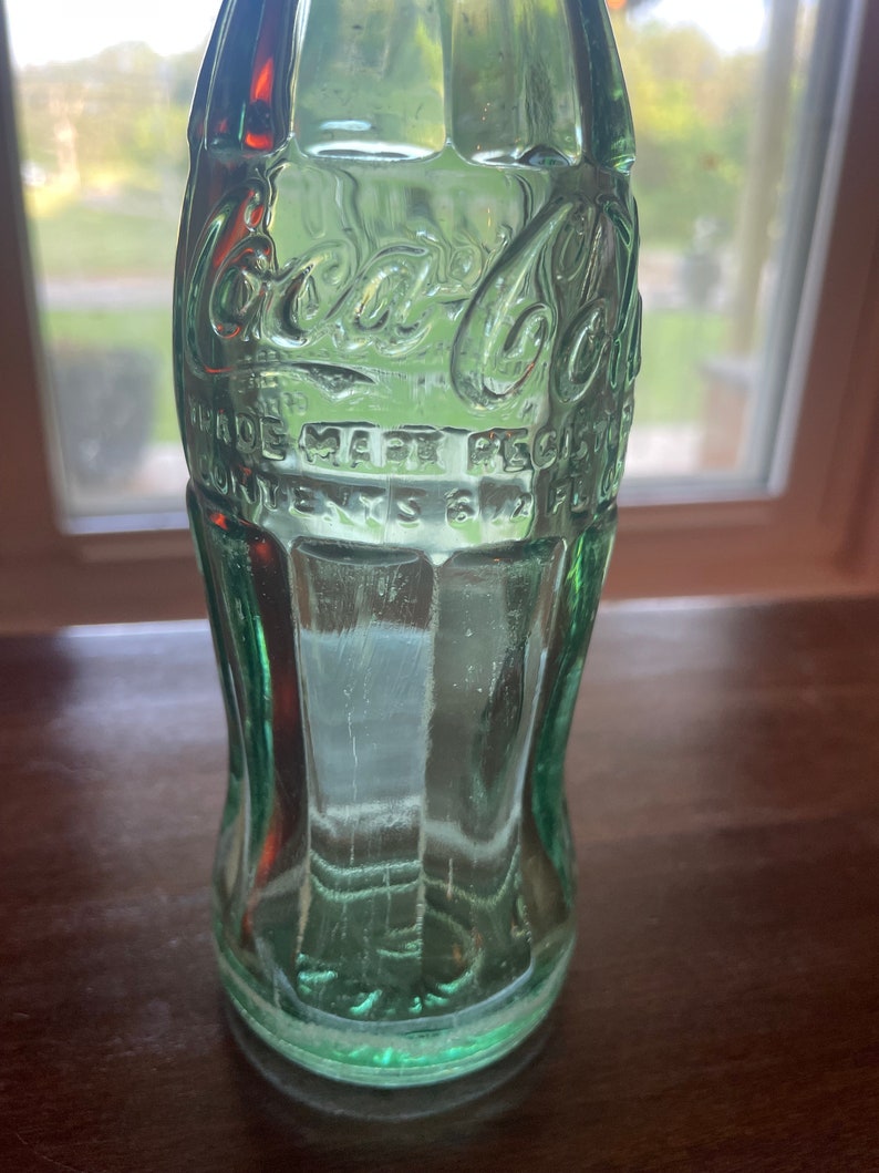 May include: A vintage green glass Coca-Cola bottle. The bottle has the Coca-Cola logo and other text embossed on the surface. The bottle is empty and has a classic, curved shape. The bottle is sitting on a dark brown surface.