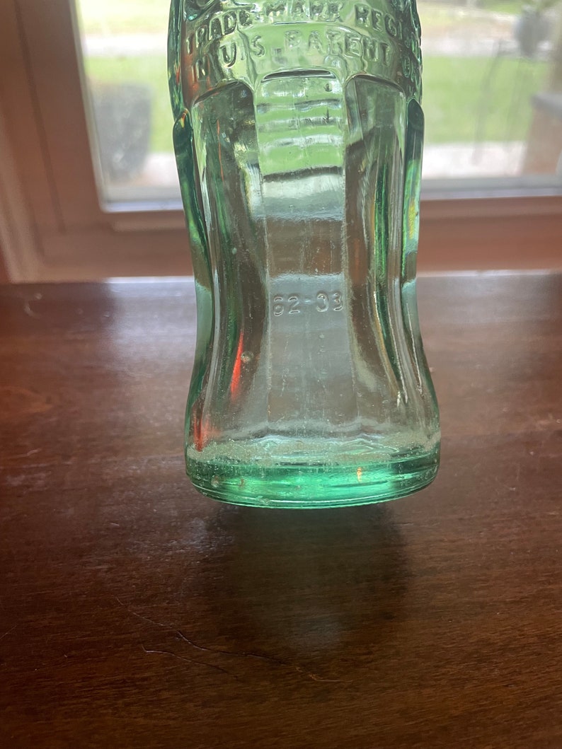 May include: A vintage green glass bottle with the text "TRADE MARK REG IN US PATENT" embossed on the upper portion. The number "62-33" is visible near the base. The bottle has a classic, curved shape.