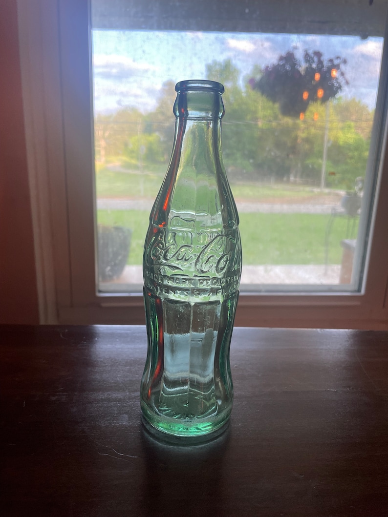 May include: A vintage green Coca-Cola bottle. The bottle is made of glass and has the Coca-Cola logo embossed on the front. The bottle is empty and sits on a dark wooden surface. The background shows a window with a view of trees and a cloudy sky.