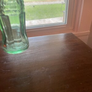 May include: A green glass bottle, partially visible, with a classic shape. The bottle is positioned on a dark brown wooden surface. The background includes a window with a view of green grass and trees.