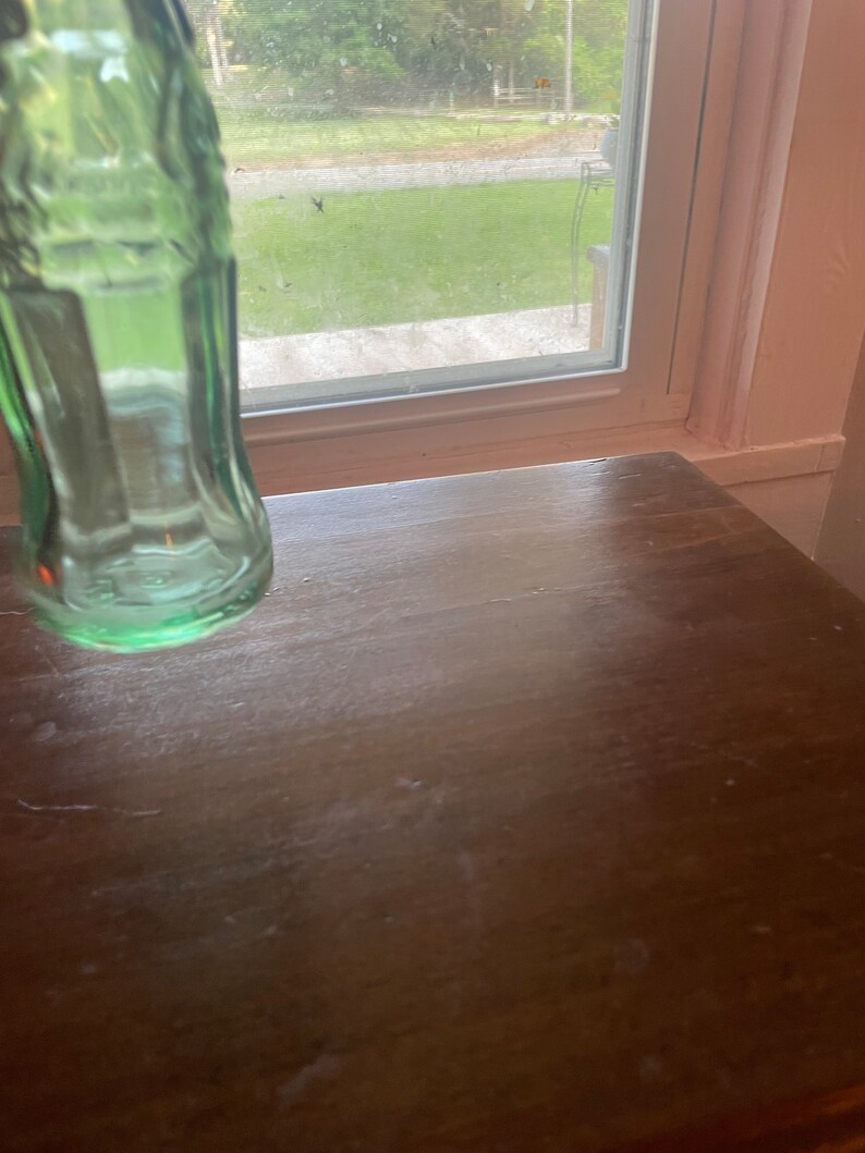 May include: A green glass bottle, partially visible, with a classic shape. The bottle is positioned on a dark brown wooden surface. The background includes a window with a view of green grass and trees.