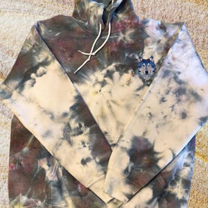 May include: A tie-dye hoodie with a wolf graphic on the front. The hoodie is a light cream color with dark blue, purple, and brown tie-dye patterns.