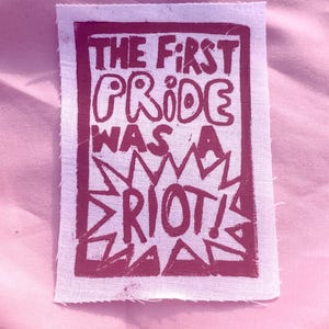 May include: A rectangular fabric patch with raw, frayed edges featuring a dark magenta screen-printed design. The text reads THE FIRST PRIDE WAS A RIOT! in a bold, hand-drawn style, enclosed within a rectangular border with a starburst graphic.