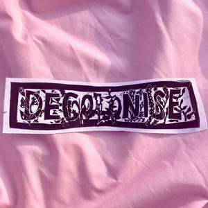 May include: A rectangular white fabric patch featuring the word DECOLONISE printed in bold black block letters. The text is enclosed within a black rectangular border, and the letters contain intricate black floral and vine-like patterns.
