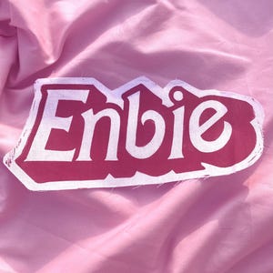 May include: A fabric patch featuring the word Enbie in white, bold, stylised lettering set against a dark pink background. The patch has a distinct, angular, and layered design with visible fabric edges and a textured, matte finish.