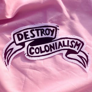 May include: A white fabric patch featuring a black screen-printed design of a two-part ribbon banner. The text "DESTROY" is printed on the top section of the banner, and the word "COLONIALISM" is printed on the bottom section of the banner.