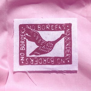 May include: A rectangular white fabric patch featuring a dark magenta screen-printed design of a bird in flight centred within a square frame. The text "NO BORDERS" is printed along all four sides of the frame in a bold, sans-serif typeface.