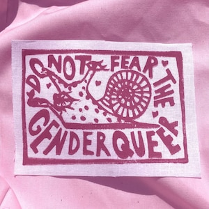 May include: A rectangular fabric patch featuring a dark pink screen-printed design on a light pink background. The graphic depicts a snail with a dotted shell and the text "DO NOT FEAR THE GENDERQUEER" arranged in a border around the central snail figure.