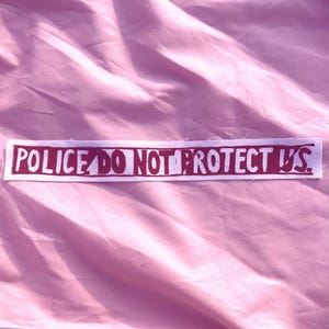 May include: A rectangular white fabric patch featuring the text POLICE DO NOT PROTECT US printed in a bold, dark red, distressed block font. The patch has raw, slightly frayed edges and is positioned horizontally against a soft pink fabric background.