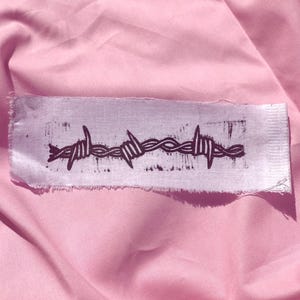 Op de afbeelding: Een rechthoekige stoffen patch met rafelige randen voorzien van een donkere, verweerde zwarte inktprint van een prikkeldraadstreng die horizontaal in het midden op het lichte, geweven textieloppervlak is geplaatst.