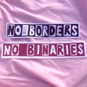 May include: Two rectangular fabric patches with raw edges. The top patch is black with white text reading NO BORDERS. The bottom patch is dark red with white text reading NO BINARIES. Both patches feature block-style lettering.