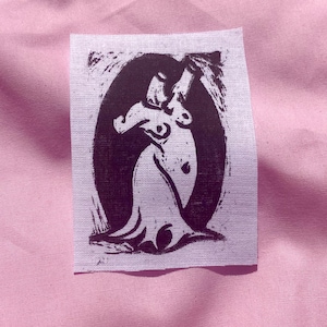 May include: A rectangular fabric patch featuring a dark purple block print illustration of a stylised figure with long hair and a flowing garment, centred within a dark oval frame on a light-coloured, textured fabric background.
