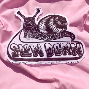 May include: A black and white fabric patch featuring a detailed linocut-style illustration of a snail with a skull-like face pattern. Below the snail, the text SLOW DOWN is printed in a bold, stylized font. The patch has a die-cut edge.