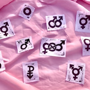May include: A collection of nine square white fabric patches featuring dark purple screen-printed gender symbols. Each patch displays a unique combination of symbols, including interlocking male, female, and non-binary icons with visible ink texture.