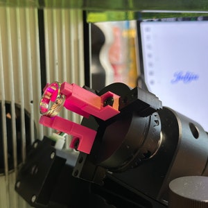 Op de afbeelding: Een gouden ring wordt gescand door een 3D-scanner. De ring wordt op zijn plaats gehouden door een roze plastic klem. De scanner is gemonteerd op een zwarte basis met een zilveren knop.