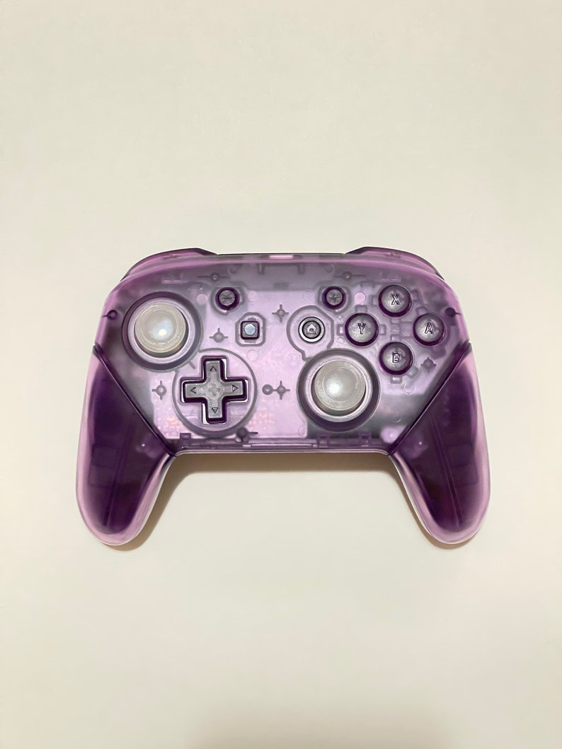 May include: A translucent purple video game controller with buttons and joysticks. The controller has a clear plastic shell and is designed for use with a gaming console.