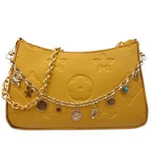 May include: A mustard yellow handbag with a textured surface featuring embossed flower and star patterns. The bag is adorned with a gold-toned chain strap that includes various small charms, including a red stone, a turquoise bead, and a round charm with the text Capricorn.
