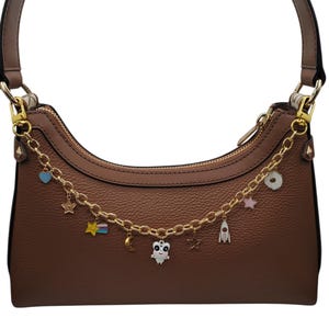 May include: A brown leather shoulder bag featuring a gold-toned chain strap adorned with various charms, including a small white alien figure, a rocket ship, a crescent moon, stars, a heart, and a colorful shooting star, all attached to the bag.