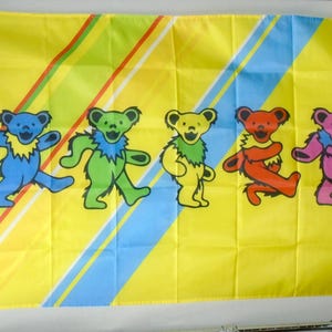 May include: A yellow flag featuring five dancing bear illustrations in blue, green, yellow, red, and pink. The bears have a zig-zag collar and are set against a background of diagonal stripes in green, red, and blue.