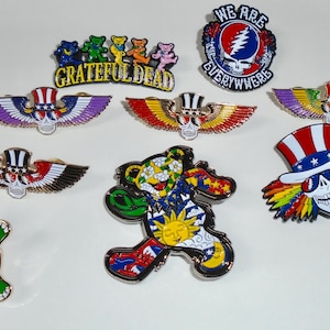 May include: A collection of ten Grateful Dead pins featuring various designs, including a skull with wings, a bear, a turtle, and a skull with a top hat. The pins are all gold-colored with colorful enamel details. The text "We Are Everywhere" is featured on one of the pins.