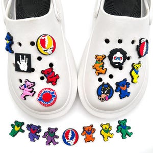 Könnte beinhalten: Weiße Clogs mit bunten, cartoonartigen Charms verziert. Die Charms zeigen verschiedene Designs, darunter tanzende Bären in verschiedenen Farben, einen Totenkopf und ein Handsymbol. Zusätzliche Charms sind unter den Clogs verstreut.