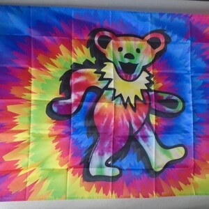 May include: A rectangular flag featuring a central graphic of a dancing bear with a black outline, set against a vibrant, multi-colored tie-dye background of blue, yellow, red, and green. The bear has a yellow ruff and a multi-colored body.