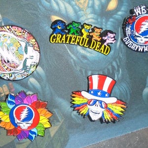 May include: A collection of colorful enamel pins featuring Grateful Dead imagery. Designs include the Steal Your Face skull, dancing bears, and Uncle Sam skull. The pins are in various shapes and sizes, with vibrant colors and intricate details.