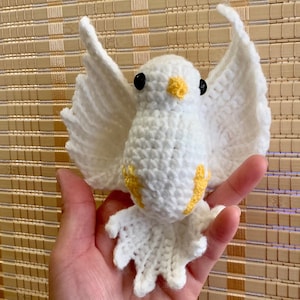 May include: A white crocheted dove with yellow accents. The dove has black eyes and a small yellow beak. The dove is being held in a person's hand.