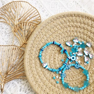 May include: A set of blue and white beaded bracelets with various charms, including rainbows, smiley faces, and evil eyes. The bracelets are displayed on a woven straw mat.