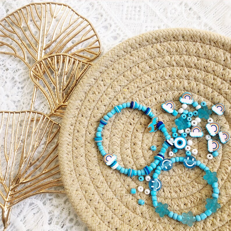 May include: A set of blue and white beaded bracelets with various charms, including rainbows, smiley faces, and evil eyes. The bracelets are displayed on a woven straw mat.