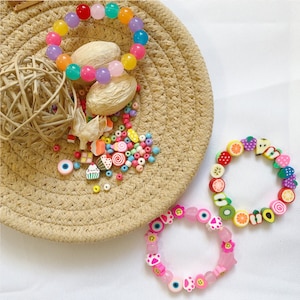 May include: Two colorful bracelets made with colorful plastic beads. One bracelet is pink with paw prints and smiley faces. The other bracelet is multi-colored with fruit shapes.