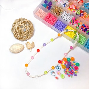 May include: A colorful necklace made with white beads and rainbow colored beads. The necklace is laid out on a white surface with a small pile of colorful beads and charms next to it.