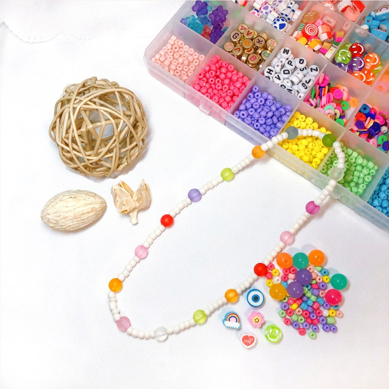 May include: A colorful necklace made with white beads and rainbow colored beads. The necklace is laid out on a white surface with a small pile of colorful beads and charms next to it.