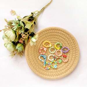 May include: A set of colorful beaded rings with various charms, including a smiley face, an eye, a heart, a paw print, and fruit. The rings are displayed on a woven straw mat.