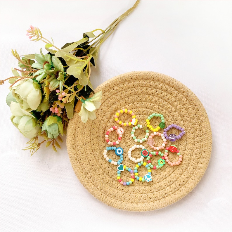 May include: A set of colorful beaded rings with various charms, including a smiley face, an eye, a heart, a paw print, and fruit. The rings are displayed on a woven straw mat.