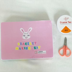 May include: A pink box with a white bunny and the text "Lucky Bunny Art Bracelet Making Kit". The box is next to a small container of clear glue labeled "Crystal Tec 0.8" and a pair of pink scissors.