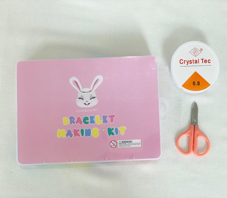 May include: A pink box with a white bunny and the text "Lucky Bunny Art Bracelet Making Kit". The box is next to a small container of clear glue labeled "Crystal Tec 0.8" and a pair of pink scissors.