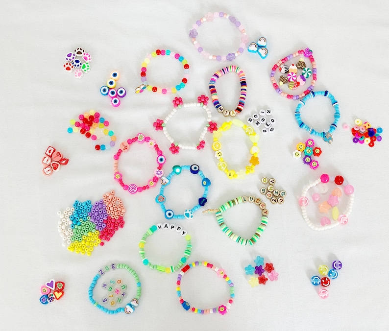 May include: A collection of colorful beaded bracelets with various charms and letters. The bracelets are made with different colored beads, including pink, blue, green, yellow, and white. Some of the charms include hearts, stars, and letters. The bracelets are arranged on a white surface.