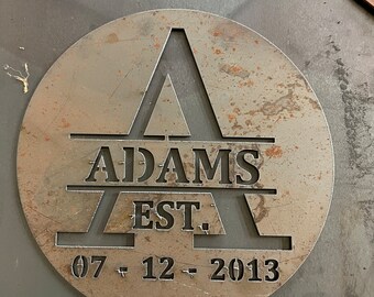 May include: A circular metal sign with the name "ADAMS" and the date "07 - 12 - 2013" cut out. The letter "A" is prominently displayed in the center. The sign has a rustic, weathered appearance.