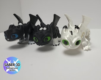 May include: Three 3D printed figurines of Toothless, the Night Fury dragon from the How to Train Your Dragon franchise. The figurines are in black, white, and a light grey colour. The figurines have green eyes and white markings.