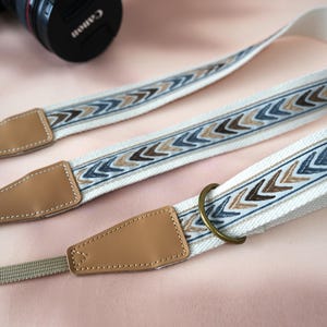 May include: Cream-colored camera strap with a geometric arrow pattern in shades of blue and brown. The strap features tan leather ends and a metal ring for attachment. A Canon camera lens is visible in the background.