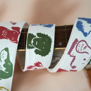 Puede incluir: Correa de guitarra blanca con ilustraciones de perros en rojo, verde, azul y morado. La correa est&aacute; envuelta alrededor del m&aacute;stil de una guitarra, mostrando el patr&oacute;n repetido de caras de perros. La correa est&aacute; hecha de un material resistente.