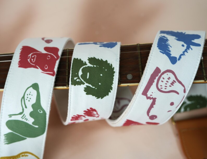 Puede incluir: Correa de guitarra blanca con ilustraciones de perros en rojo, verde, azul y morado. La correa est&aacute; envuelta alrededor del m&aacute;stil de una guitarra, mostrando el patr&oacute;n repetido de caras de perros. La correa est&aacute; hecha de un material resistente.