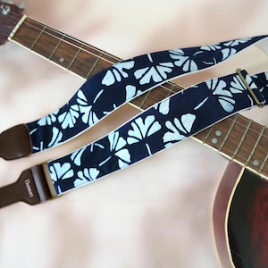 May include: A navy blue and white floral guitar strap with brown leather ends and a metal buckle. The strap is draped over a brown and wooden guitar.